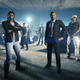 A woman in a leather jacket and a man in a black business suit stand in front of a large group of GTA Online players.